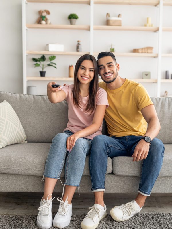 Joyful diverse couple watching TV program, sitting on couch at home, full length portrait